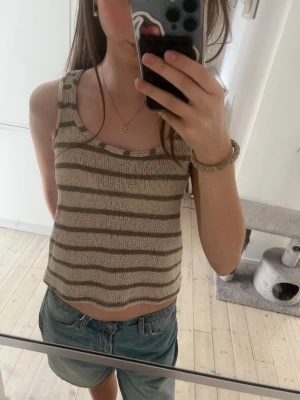Beige och brun randig stickad linnetopp - Säljer en stickad linnetopp i beige och brunt med horisontella ränder. Toppen är ärmlös med breda axelband och har en croppad passform. Perfekt till jeans eller kjol för en chill look.