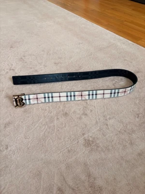 Rutmönstrat bälte från Burberry - Snyggt bälte från Burberry med klassiskt rutmönster i vitt, svart och rött. Bältet har ett metallspänne med Burberrys logotyp och är gjort i skinn. Perfekt accessoar för att lyfta din outfit.