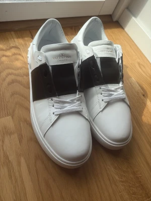 Valentino vita sneakers med svart detalj - Snygga vita sneakers från Valentino Garavani med bred svart läderdetalj över snörningen och coola nitar längs hälen. Skorna har klassisk låg profil, vita skosnören och en stilren design som passar till det mesta. Perfekta för dig som gillar exklusiva märken och streetstyle.