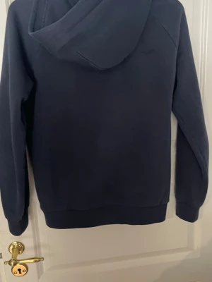 Mörkblå hoodie Peak Performance - Mörkblå hoodie från Peak Performance med vit logga på bröstet och klassisk känguruficka framtill. Hoodien har ribbade muddar vid ärmslut och nederkant samt en stor huva. Perfekt för en avslappnad och sportig stil.