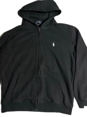 Svart hoodie från Polo Ralph Lauren - Klassisk svart hoodie från Polo Ralph Lauren med dragkedja framtill och vit broderad logga på bröstet. Modellen har huva, kängurufickor och ribbade muddar. Tillverkad i mjuk bomullsblandning för skön komfort. Perfekt till chill eller streetwear-stil.