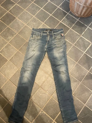 Blå slim fit jeans från Replay - Snygga blå slim fit jeans från Replay med slitningar och tvättade detaljer. Klassisk femficksmodell med dragkedja och knapp i midjan. Jeansen har en skön stretch och Replay-logga på bakfickan. Perfekta för en avslappnad och trendig look.