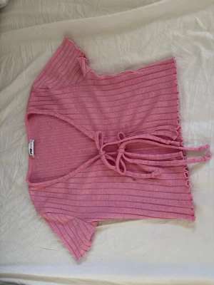 Rosa ribbad knytblus - Söt rosa ribbad topp med korta ärmar och knyt framtill. Toppen har en v-ringad urringning och vågiga kanter vid ärmslut och nederkant. Perfekt för en trendig och avslappnad look.
