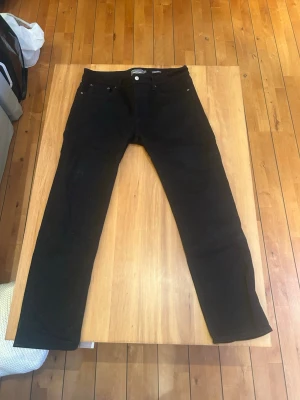 Svarta slim fit jeans från Dressman - Säljer ett par svarta slim fit jeans från Dressman. Klassisk femficksmodell med knappgylf och raka ben. Jeansen är tillverkade i stretchigt denimtyg för extra komfort och har en stilren look som passar till det mesta.