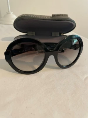 Svarta runda solglasögon från Prada - Snygga svarta solglasögon från Prada med rund oversized form och mörka glas. Klassisk och trendig design med diskret Prada-logga på skalmen. Perfekta för dig som vill sticka ut med en cool accessoar.