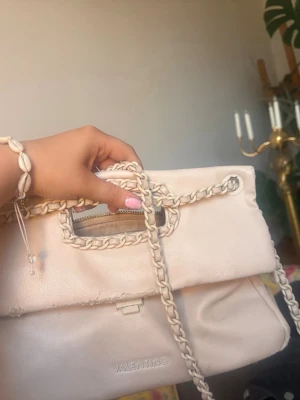 Ljusbeige handväska från Valentino - Snygg ljusbeige eller ljusrosa / rosa handväska från Valentino med flätade handtag och axelrem. Väskan har ett stilrent och mjukt material med präglad logga framtill. Perfekt för dig som vill ha en trendig och elegant accessoar till din outfit. Bandet går att justera så att man får en axelväska eller en över axeln. Rymmer mycket 