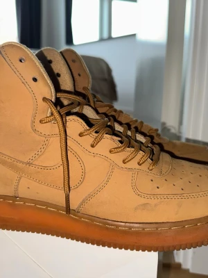 Nike Air Force 1 High Wheat - Snygga Nike Air Force 1 High i färgen wheat. Skorna är tillverkade i mocka med matchande bruna skosnören och en gummisula. Klassisk high-top siluett med perforerad tåbox och ikoniska swoosh-detaljer på sidorna. Perfekta för dig som gillar streetwear och stilrena sneakers.