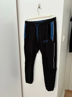 Svarta mjukisbyxor med blå detaljer - Säljer ett par svarta mjukisbyxor från Hugo boss byxor från deras gamla collektion. Urban med blå detaljer vid fickorna och blå dragsko i midjan. Byxorna har smal passform, vita ränder längs ena benet och tryckt logga på vänster sida. Perfekta för chill eller träning.