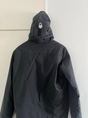 Svart vindjacka från Sail Racing - Snygg svart vindjacka från Sail Racing med huva och hel dragkedja framtill. Jackan har en minimalistisk design, diskret logga på bröstet och justerbar huva med kardborreband. Tillverkad i lätt och vindtätt material, perfekt för blåsiga dagar. Storlek S, inte använd så mycket. Bra skick även.