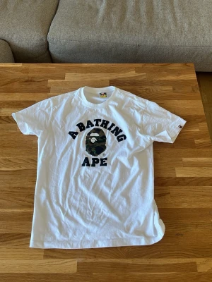 Vit A Bathing Ape t-shirt med camo - Säljer en vit t-shirt från A Bathing Ape med klassiskt camo-tryck av apans ansikte och stor svart text på bröstet. T-shirten är kortärmad och har normal passform. Tillverkad i mjuk bomull, perfekt för dig som gillar streetwear och vill sticka ut.