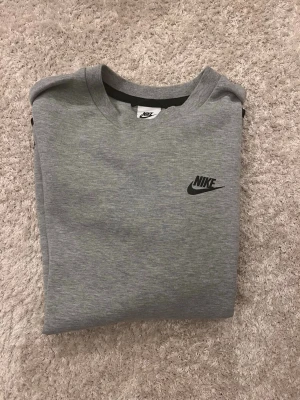 Grå nike tech sweatshirt - Snygg grå Nike tech  sweatshirt med svart logga på bröstet och en cool ficka med dragkedja på ena ärmen. Tröjan har rund halsringning och svarta detaljer vid ärmslut och nederkant. Perfekt för en sportig och avslappnad stil.