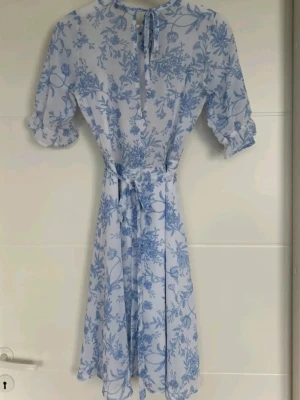 Blåvit blommig kortklänning med knyt☀️  - Söt kortklänning i vitt med blått blommönster🌸 Klänningen har halvlånga puffärmar med volangkant, knytband i nacken och ett matchande midjeband🩵 Tillverkad i ett lätt och luftigt material, perfekt för varma dagar😍☀️ XS-S. 