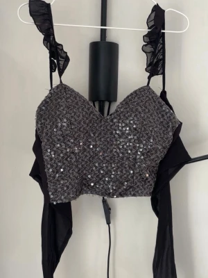 Svart paljett korsettopp med volang - Trendig svart korsettopp med paljetter framtill och volangdetaljer på axelbanden. Toppen har omlottband i mesh som knyts i midjan och en hjärtformad urringning. Perfekt för dig som vill sticka ut med en glittrig och unik topp. Aldrig använd.