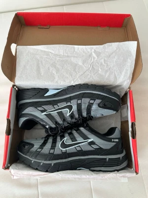 Nike P-6000 svart/grå sneakers - Nike P-6000 sneakers i svart och grått med vit swoosh. Skorna har en sportig design med mesh och syntetiska överdrag, samt robust sula för extra komfort. Perfekta för dig som gillar streetwear och vill ha en cool, modern look.