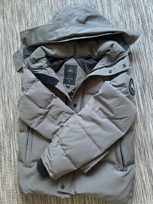 Grå canada goose jacka - Snygg grå dunjacka från Canada Goose med huva och diskret logotyp på ärmen. Jackan har dragkedja och knappar framtill, två sidofickor med dragkedja och ribbade muddar vid ärmsluten. Perfekt för kalla vinterdagar.