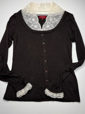 Brun blus med spets från Burberry - Stilren brun blus från Burberry med lång ärm, dekorativa knappar framtill och vit spetskrage med broderade detaljer. Blusen har även spetsdetaljer vid ärmsluten och nederkanten. Tillverkad i mjukt tyg som ger en elegant look. Perfekt för dig som gillar klassiska och unika plagg.