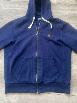 Marinblå hoodie från Polo Ralph Lauren - Snygg marinblå hoodie med dragkedja från Polo Ralph Lauren. Klassisk gul logga broderad på bröstet, vita dragsnören i huvan och känguruficka framtill. Tillverkad i mjuk bomullsblandning som känns skön mot huden. Perfekt för en avslappnad och stilren look.