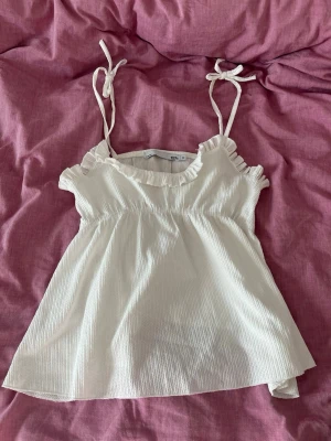 Vit linnetopp med volang  - Supersöt vit linnetopp med volangdetaljer vid ringningen och smala axelband som knyts upptill. Storlek M men passar som S också