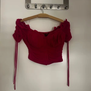 Röd offshoulder croppad topp - Trendig röd offshoulder topp med croppad passform och långa knytband vid ärmarna. Toppen har rynkade detaljer framtill och en feminin siluett som passar perfekt till höga jeans eller kjol.