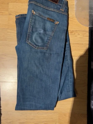 Blå jeans från Nudie Jeans - Säljer ett par klassiska blå jeans från Nudie Jeans med raka ben och fem fickor. Jeansen har snygga kontrastsömmar och en patch bak i midjan. Tillverkade i mjuk denim som sitter skönt och passar till det mesta. Perfekta för dig som gillar stilrena och tidlösa jeans.