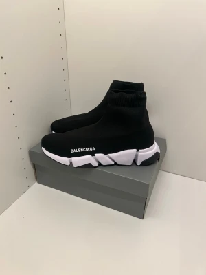 Svart vita b skor  - Svarta B skor sula och stickad strumpliknande ovandel. Minimalistisk design med logga på sidan och elastisk passform som sitter tajt runt ankeln. Perfekta för dig som gillar streetwear och modern stil.