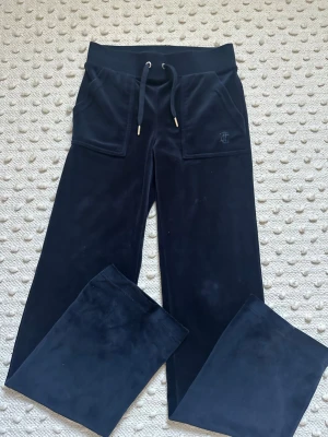 Mörkblå velourbyxor Juicy Couture - Säljer ett par mörkblå mjukisbyxor från Juicy Couture i mjuk velour. Byxorna har breda ben, dragsko i midjan, stora fickor fram och bak med guldfärgade knappar samt broderad logga fram. Perfekta för en chill och trendig look.
