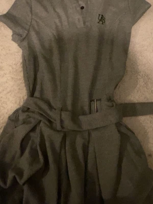 Grå ribbad klänning med bälte - Snygg grå ribbad klänning med korta ärmar och ett matchande bälte i midjan. Klänningen har en liten logga på bröstet och knappar vid halsen. Perfekt för dig som gillar en sportig och stilren look.