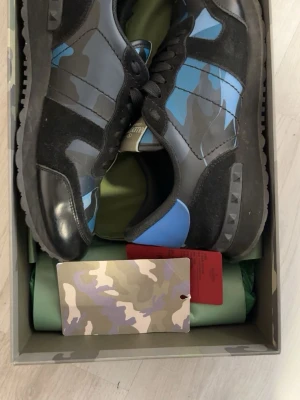 Valentino rockrunners sneakers i blått och svart - Snygga sneakers från Valentino med coolt camo-mönster i blått, svart och grått. Skorna har svarta mockadetaljer, läderpartier och en blå accent på hälen. Yttersulan har nitar och ger ett edgy uttryck. Perfekt för dig som vill sticka ut med stil.