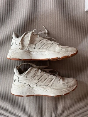 Beige Adidas sneakers med gummisula - Snygga beige sneakers från Adidas med klassiska tre ränder på sidan och detaljer i mocka och mesh. Skorna har en chunky vit mellansula och yttersula i gummi för extra grepp. Perfekta för dig som gillar en sportig och trendig look.