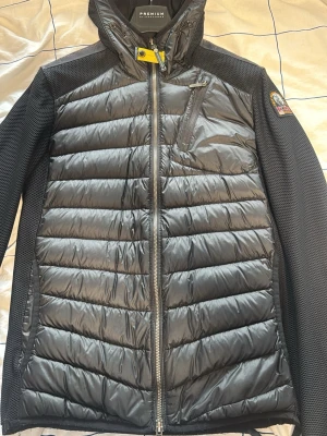 Svart pufferjacka från Parajumpers - Svart pufferjacka från Parajumpers med quiltad front och huva. Jackan har en bröstficka med dragkedja, mesh-paneler på sidorna och ärmarna samt ett karakteristiskt gult spänne vid halsen. Märkespatch på vänster ärm. Perfekt för kalla vinterdagar.