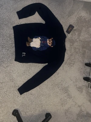 Mörkblå stickad Ralph lauren tröja med björn motiv - Säljer en mörkblå stickad tröja från Polo Ralph Lauren med deras ikoniska björnmotiv på bröstet. Tröjan har långa ärmar, ribbade muddar och RL-broderi nedtill. Perfekt för dig som gillar klassiska och lekfulla detaljer.