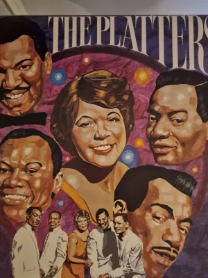 Vinylskiva The Platters - Vinylskiva med omslag av The Platters. Omslaget har färgglad illustration av bandmedlemmarna mot en lila bakgrund med färgglada cirklar. Baksidan listar låtar och information om bandet. Perfekt för samlare av klassisk musik.