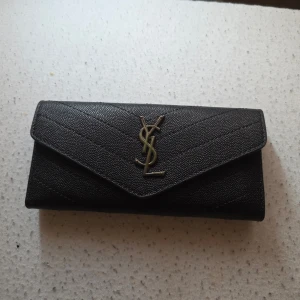 Svart YSL plånbok i läder - Snygg svart plånbok från YSL i äkta läder med guldfärgad logga framtill. Plånboken har flera kortfack, ett fönster för ID och ett större fack för sedlar. Klassisk kuvertformad design som känns både lyxig och tidlös.