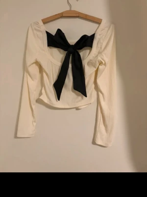 Vit långärmad topp med svart rosett - Superfin vit långärmad topp med öppen rygg och stor svart rosett i ryggen. Toppen är croppad och har en snygg, figurnära passform. Oanvänd utan prislapp
