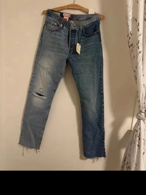 Levi's blå slitna jeans med hål - Säljer ett par blå Levi's jeans med rak passform, slitningar och hål på ena benet samt råa, fransiga benslut. Klassisk femficksmodell i denim med vintagekänsla. Helt oanvända! 28x32