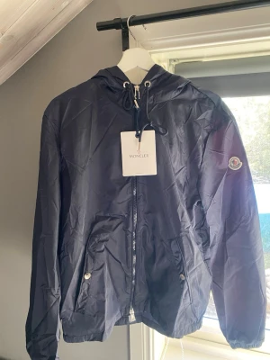 Mörkblå Moncler vindjacka med huva - Snygg mörkblå vindjacka från Moncler med huva och dragkedja framtill. Jackan har två fickor med knappar och Moncler-logga på ärmen. Materialet är lätt och vattenavvisande, perfekt för blåsiga dagar. Huva med röd, vit och blå detalj baktill.