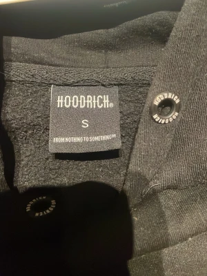 Svart hoodie från Hoodrich - Svart hoodie från Hoodrich med stor vit logga broderad över bröstet. Klassisk känguruficka framtill och justerbar huva med snören. Mjuk insida och ribbade muddar vid ärmslut och nederkant. Perfekt för en streetwear-look.