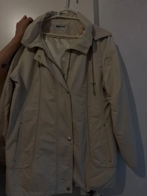 Beige vindjacka från Almanda - Stilren beige vindjacka från Almanda med dragkedja och tryckknappar framtill. Jackan har två stora fickor med dragkedja, hög krage och justerbar dragsko nertill. Perfekt för blåsiga dagar och enkel att matcha med olika outfits.