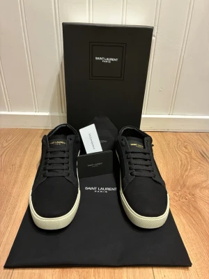 Saint Laurent sneakers - Svarta sneakers från Saint Laurent Paris med vit sula och svart ovandel i canvas. Snyggt broderad Saint Laurent-logga på sidan och guldfärgad text på plösen. Klassisk låg modell med svarta skosnören och diskret design som passar till det mesta.