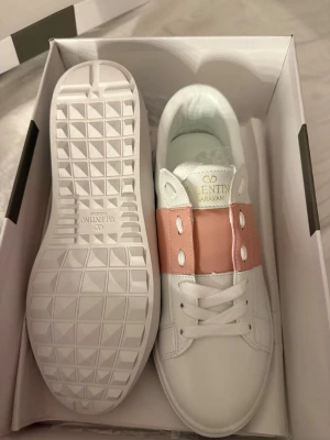 Rosa Valentino Garavani - Snygga vita sneakers från Valentino Garavani med bred ljusrosa läderdetalj över snörningen och guldfärgad logga på plösen. Skorna har en chunky vit sula med rutmönster undertill och är tillverkade i slätt läder. Perfekta för dig som gillar exklusiv och trendig stil.