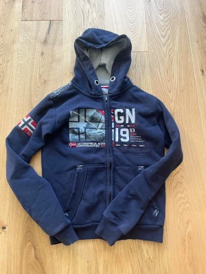 Blå hoodie från Geographical Norway - Säljer en mörkblå hoodie från Geographical Norway med coola tryck och broderade detaljer på bröstet och ärmen. Hoodien har dragkedja, två fickor och en skön huva med grått innerfoder. Perfekt för dig som gillar sportig och avslappnad stil.