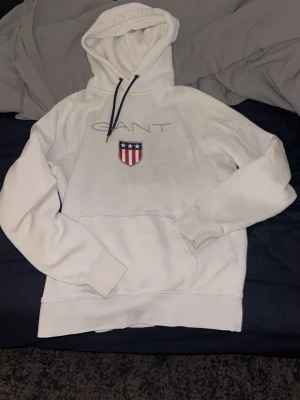 Vit hoodie GANT - Säljer en vit hoodie från GANT med stor logga och amerikanskinspirerat emblem på bröstet. Hoodien har huva med mörka dragsnören och en stor magficka. Tillverkad i mjukt bomullsmaterial, perfekt för en avslappnad och stilren look.