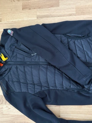 Svart pufferjacka från Parajumpers - Snygg svart pufferjacka från Parajumpers med quiltad front och rygg, samt släta ärmar och sidor. Jackan har dragkedja framtill, bröstficka med dragkedja och klassisk Parajumpers-logga på axeln. Perfekt för kalla vinterdagar.
