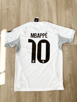 Real Madrid matchtröja Mbappé #10 - Säljer en vit Real Madrid matchtröja från Adidas med Mbappé och nummer 10 på ryggen. Tröjan har svarta detaljer, klubbmärke på bröstet, sponsortryck och gula sömmar. Tillverkad i lätt och ventilerande material, perfekt för fotbollsträning eller match.