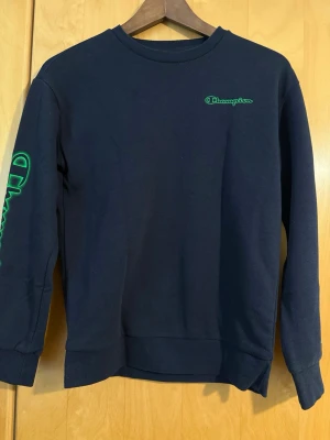  Champion tjocktröja  - Mörkblå tröja  från Champion med grön logga på bröstet och stort Champion-tryck längs ena ärmen.  10-11 år 