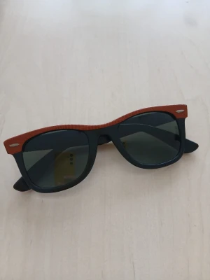  Ray-Ban Wayfarer solglasögon svart/brun - Ray-Ban snygga Wayfarer solglasögon med svart båge och brun detalj upptill. Klassisk fyrkantig form och mörka glas. Perfekta för att lyfta din stil med en retrovibe. Tillverkade i plast för en lätt känsla.