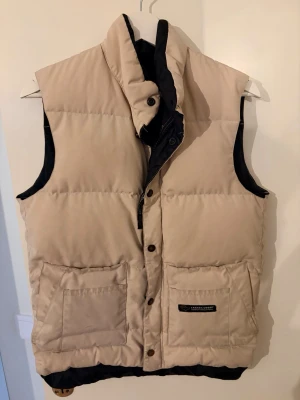 Beige dunväst från Canada Goose - Snygg beige dunväst från Canada Goose med hög krage och svarta detaljer på insidan. Västen har två stora framfickor och logotyp på ena fickan. Perfekt för lager-på-lager och håller dig varm under kyliga dagar.