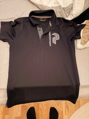 Svart pikétröja från Peak Performance - Säljer en svart pikétröja från Peak Performance med klassisk krage och knappar framtill. Tröjan har en stor logga och text på bröstet. Perfekt för en sportig och stilren look.