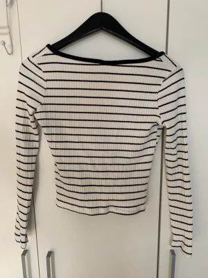Randig ribbad långärmad topp - Säljer en vit och svart randig ribbad topp med lång ärm och v-ringad hals. Toppen är croppad och har en snygg passform som framhäver figuren. Perfekt för att skapa en chill och trendig look.