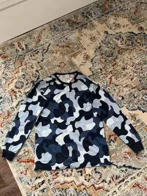 Blå camo långärmad tröja - Säljer en långärmad tröja med blått camouflagemönster i olika nyanser av blått, svart och vitt. Tröjan har svarta muddar vid ärmslut och halsringning. Perfekt för dig som gillar streetwear och vill sticka ut med en cool look.